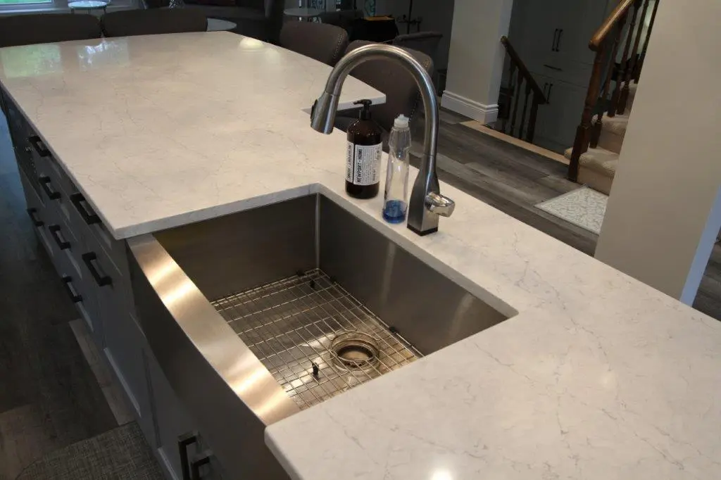 Modern kitchen island with a large stainless steel farmhouse sink, marble countertop, stainless steel gooseneck faucet, and two soap dispensers, with dark wood cabinets and chairs in the background.