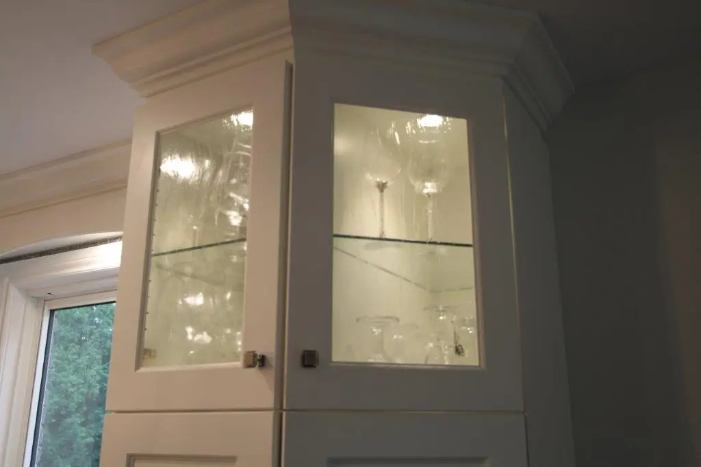 Upper section of a white kitchen cabinet with glass-paneled doors displaying several wine glasses inside, with a window and greenery visible to the left.