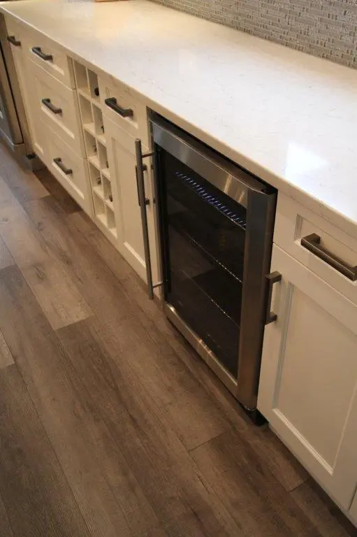 Close-up view of a kitchen counter with white cabinets, a built-in stainless steel wine cooler with a glass door, and a light-colored marble countertop, set against a stone tile backsplash and wood plank flooring.