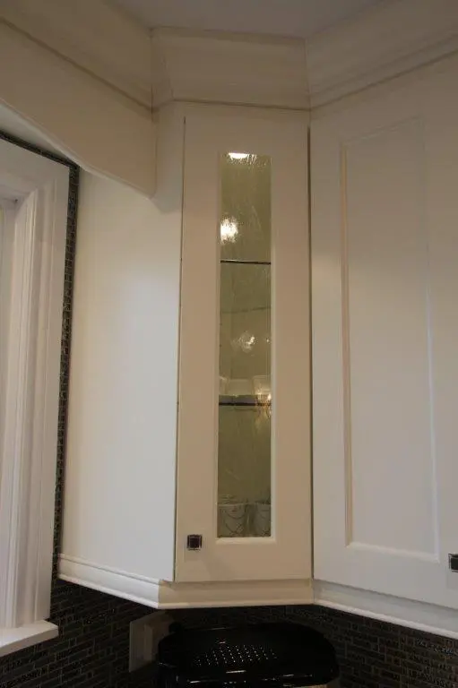 Corner section of white kitchen cabinets with a tall narrow glass panel door revealing glass shelves inside and a black appliance below, set against a dark tiled backsplash and white walls.
