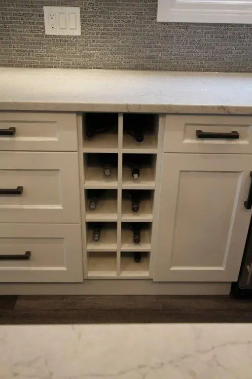 Built-in kitchen cabinet with a vertical wine rack holding eight wine bottles, surrounded by white drawers and cabinets with black handles, a light countertop above, and a gray tiled backsplash with a power outlet.