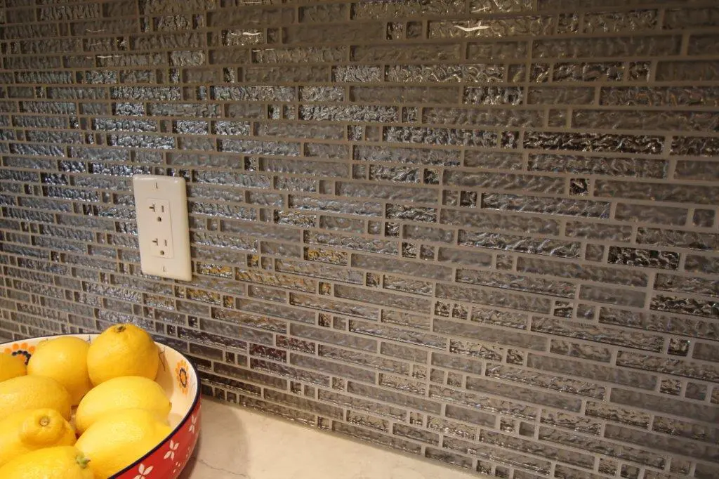 Kitchen wall with reflective gray mosaic tiles, a white electrical outlet, and a bowl of yellow lemons on a beige countertop