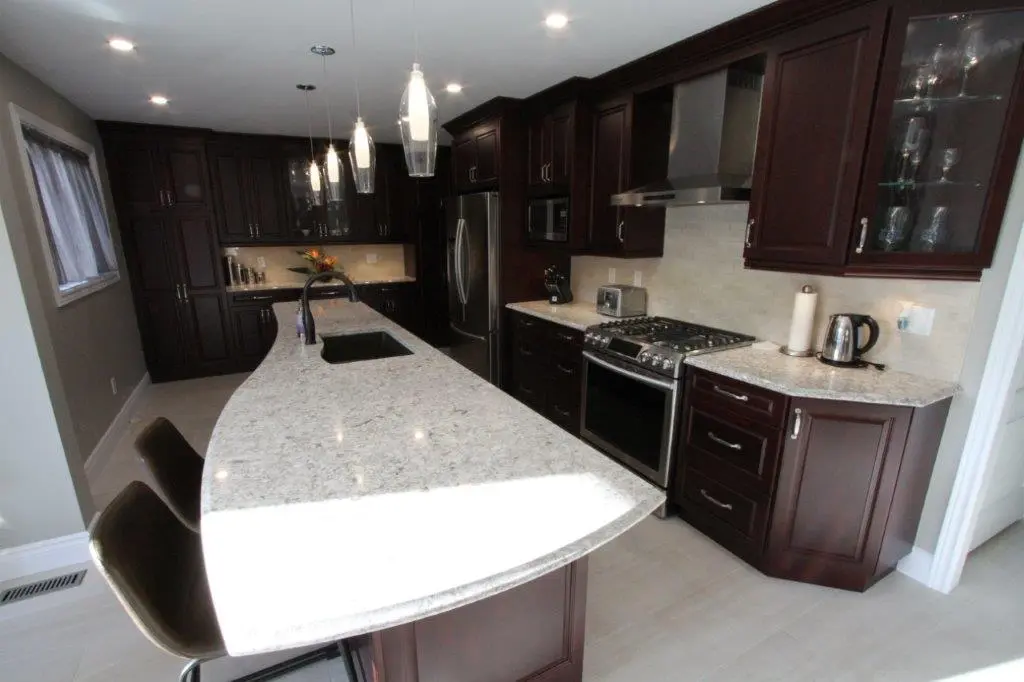 Modern kitchen with dark wood cabinets, a curved light granite countertop island with sink, three pendant lights above, stainless steel appliances including a refrigerator and gas stove, and two bar stools.