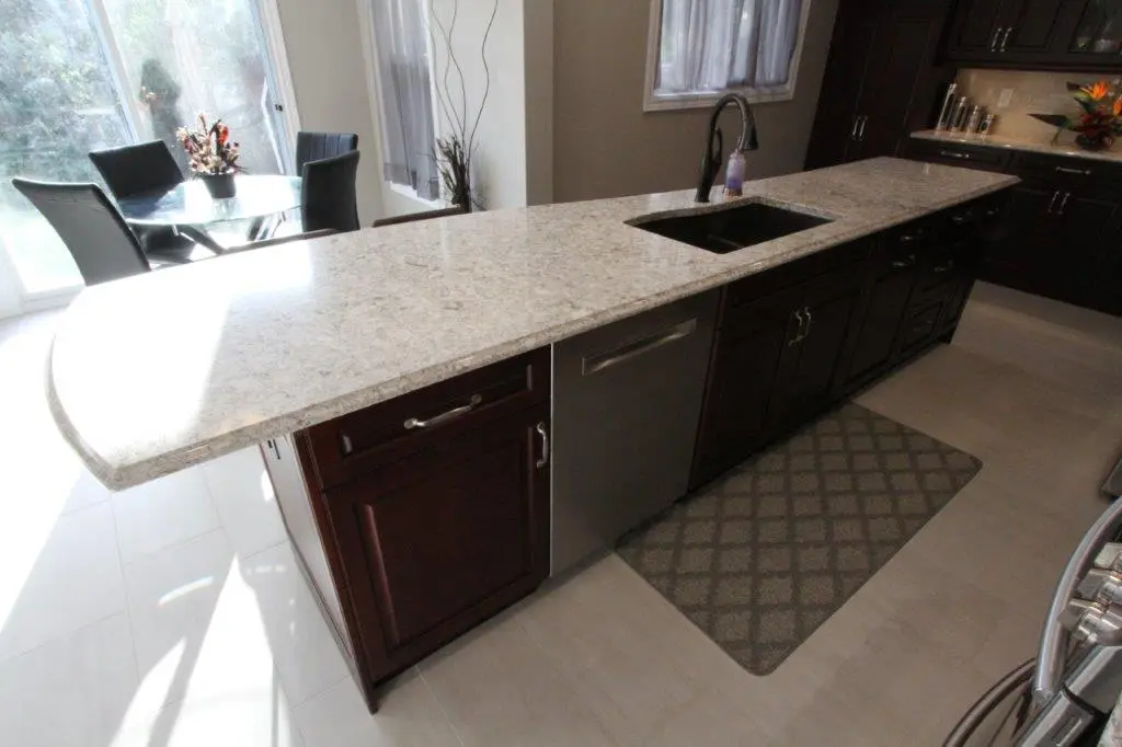 Modern kitchen island with dark wood cabinets, quartz countertop, built-in sink, and stainless steel dishwasher, with a patterned floor mat underneath and a dining area with black chairs and a glass table in the background.