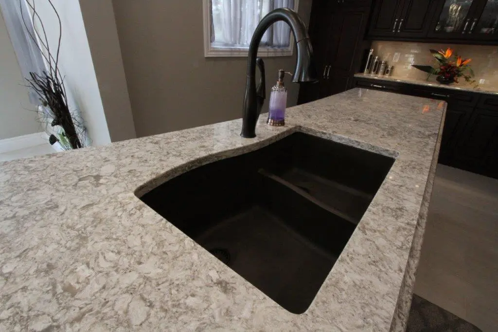 Modern kitchen countertop with a light gray and white marbled pattern, featuring a large black undermount sink and a curved black faucet with a purple soap dispenser next to it.