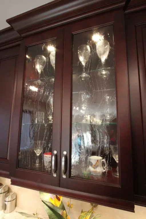 Dark wood kitchen cabinet with glass doors containing wine glasses and a mug, glass panels appear streaked or fogged
