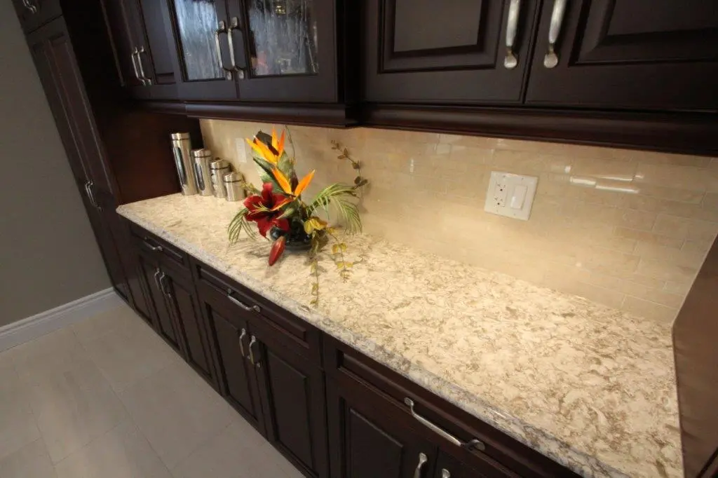 Kitchen countertop with a granite surface featuring a colorful floral arrangement in the center and stainless steel canisters on the left, surrounded by dark wooden cabinets and a beige tiled backsplash with a light switch and electrical outlet.