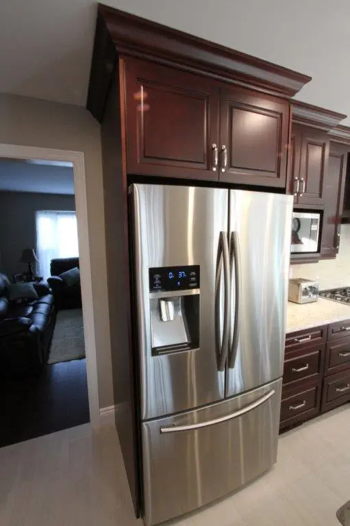 Stainless steel French door refrigerator with digital display and water dispenser set within dark wood cabinetry in a modern kitchen setting with adjacent dark wood cabinets and light-colored floor tiles.