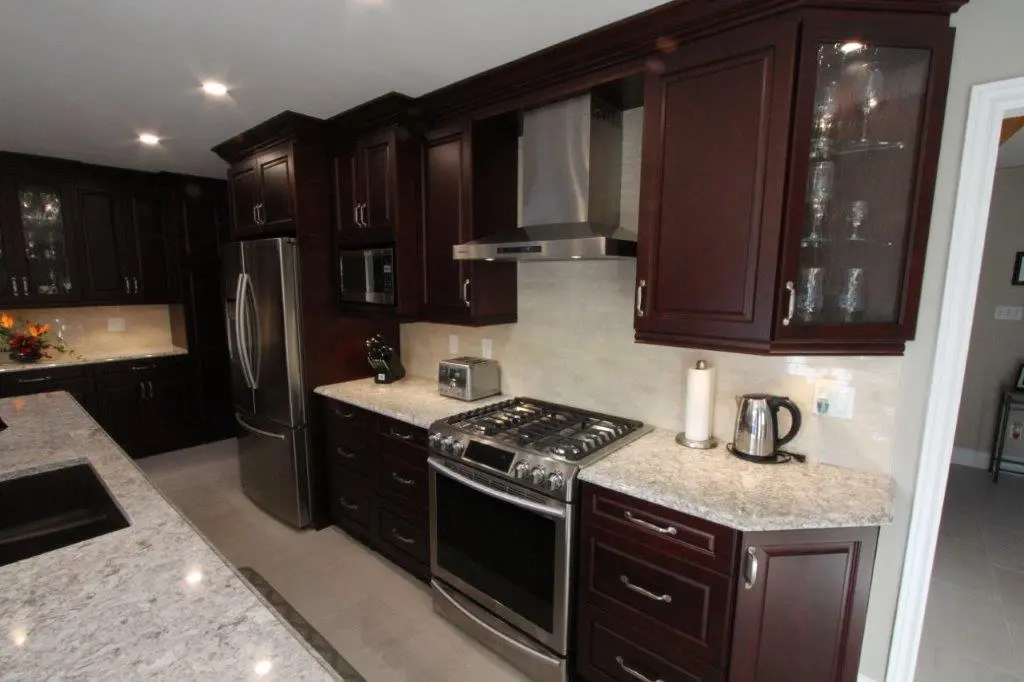 Modern kitchen with dark wood cabinets, stainless steel refrigerator and gas stove, marble countertops, and glass-front cabinet displaying glassware
