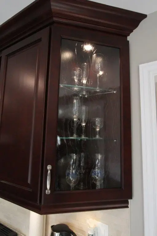 Dark wood kitchen cabinet with a glass door displaying various glassware including wine glasses and goblets on three glass shelves.
