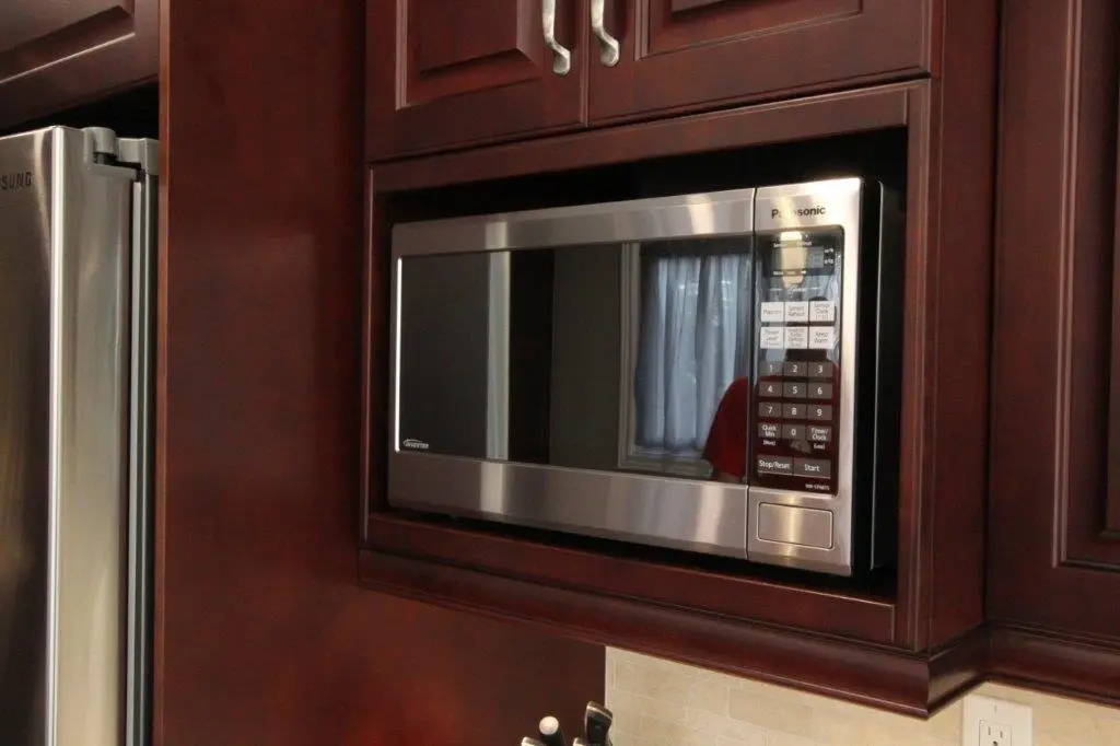 Built-in stainless steel Panasonic microwave installed in dark wooden kitchen cabinetry next to a stainless steel refrigerator.