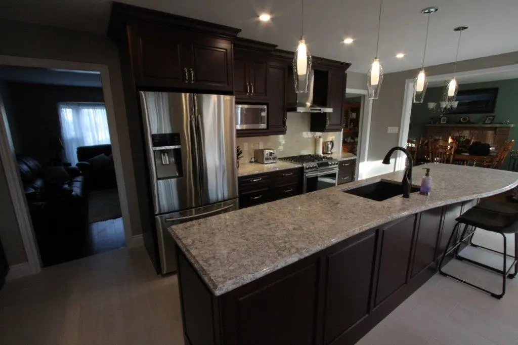 Modern kitchen with dark wood cabinets, a large granite countertop island with a built-in sink and black faucet, stainless steel refrigerator and appliances, pendant lights hanging above the island, and a glimpse into adjacent living and dining areas.