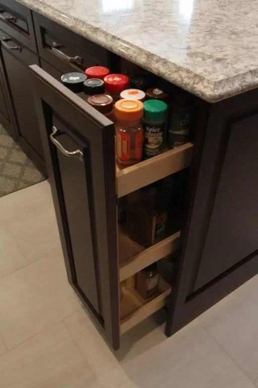 Narrow kitchen cabinet pull-out drawer filled with various spice jars under a marble countertop