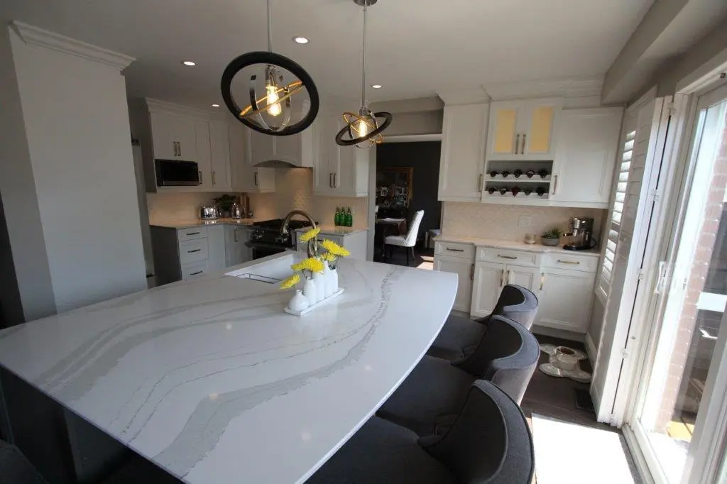 Modern kitchen with a large white quartz island countertop, dark gray upholstered bar stools, white cabinets, built-in wine rack, and two black and gold pendant lights hanging over the island, with glass doors leading outside on the right side.