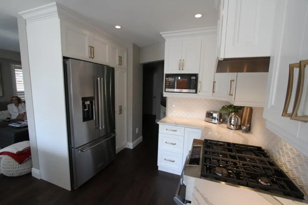 Modern kitchen with stainless steel French door refrigerator, white cabinetry, dark hardwood floors, a microwave above the counter, a gas stove with multiple burners, and small appliances on the countertop including a toaster and kettle, with an adjacent living room area.