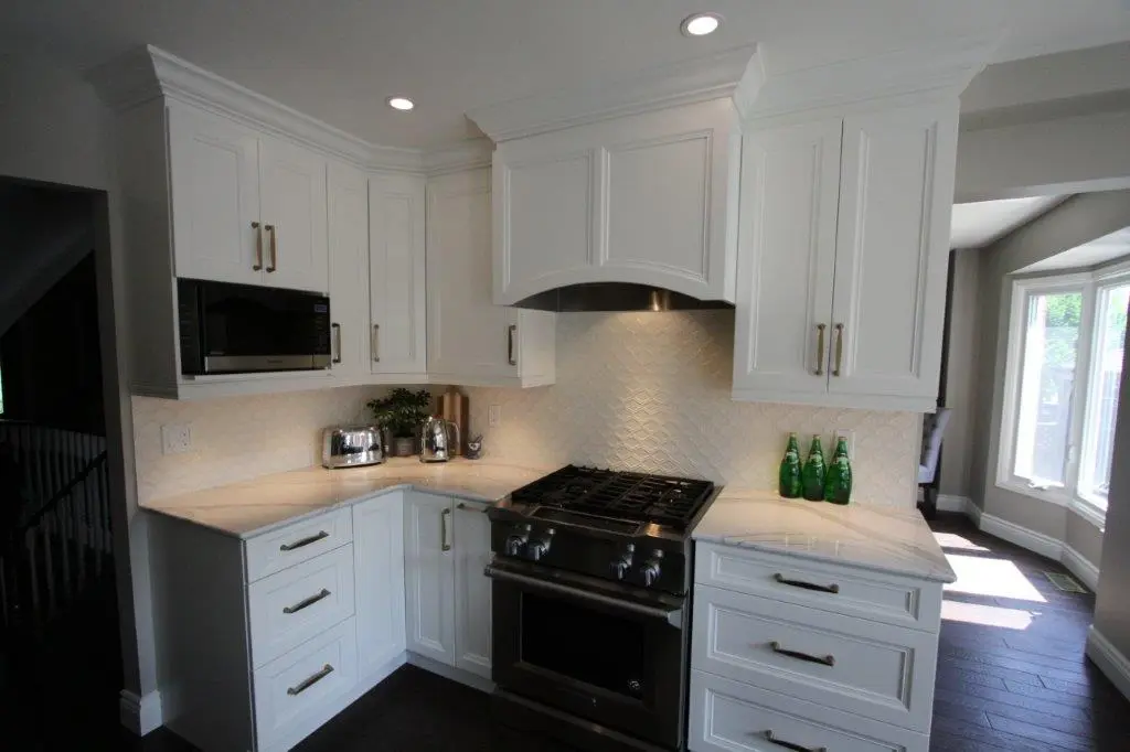 Modern kitchen corner with white cabinetry, stainless steel gas stove and oven, white quartz countertops, a built-in microwave, toaster, kettle, potted plant, and three green glass bottles, with natural light coming through a nearby window.