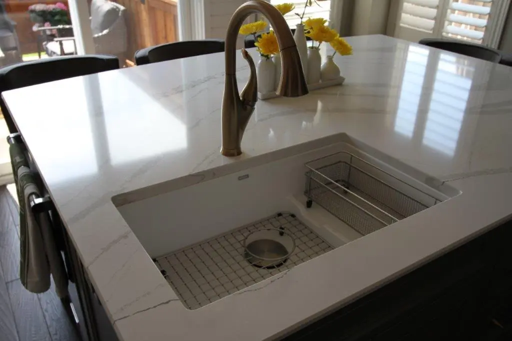 Modern kitchen island with white quartz countertop featuring an undermount sink with a bronze faucet, a metal grid at the bottom of the sink, and a wire basket, with yellow flowers in white vases placed on the countertop.
