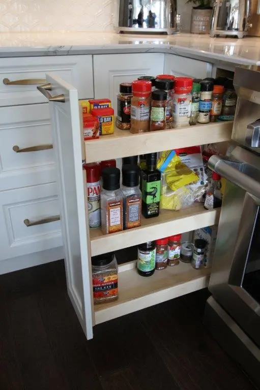 Pull-out kitchen cabinet organizer with three wooden shelves holding various spice jars, seasoning containers, and cooking ingredients next to a white cabinet and stainless steel oven.