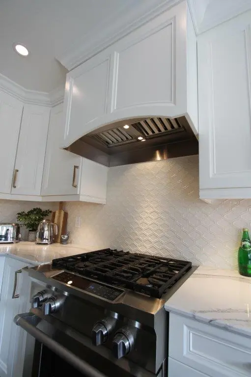 Modern kitchen with a stainless steel gas stove featuring four burners and a stainless steel range hood, surrounded by white cabinetry and a textured white backsplash, with a marble countertop holding a plant, toaster, kettle, and a green glass bottle.
