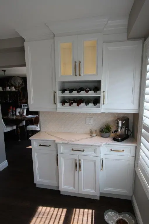 White kitchen cabinetry with a built-in wine rack holding multiple wine bottles, a small coffee machine on the right countertop, and a decorative plant next to it, set against a light tiled backsplash with an electrical outlet, with a window featuring white shutters on the right side and dark wooden flooring below the cabinets.
