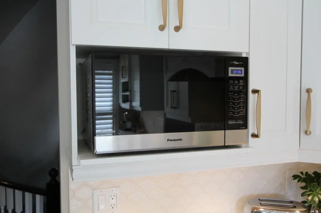 Stainless steel Panasonic microwave oven installed in white kitchen cabinetry with a digital display showing 3:45 and gold-colored handles on the cabinets.