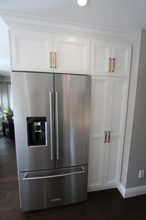Stainless steel French door refrigerator with a built-in water and ice dispenser, integrated into white cabinetry with brass handles, set against a dark wood floor in a modern kitchen.