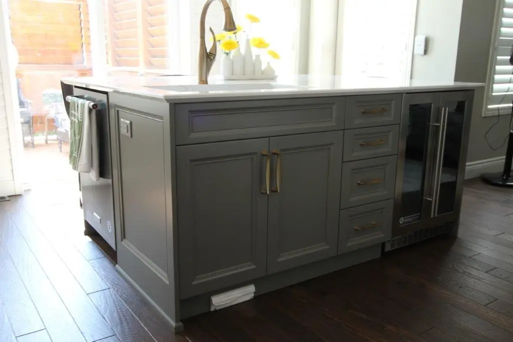Gray kitchen island with gold handles, a white countertop, a brushed gold faucet, a vase with yellow flowers, a built-in wine fridge with glass doors, and dark hardwood flooring surrounding it.