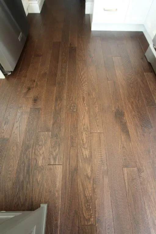 Close-up view of dark brown wooden floor planks in a kitchen with white cabinets and a stainless steel appliance visible on the left side