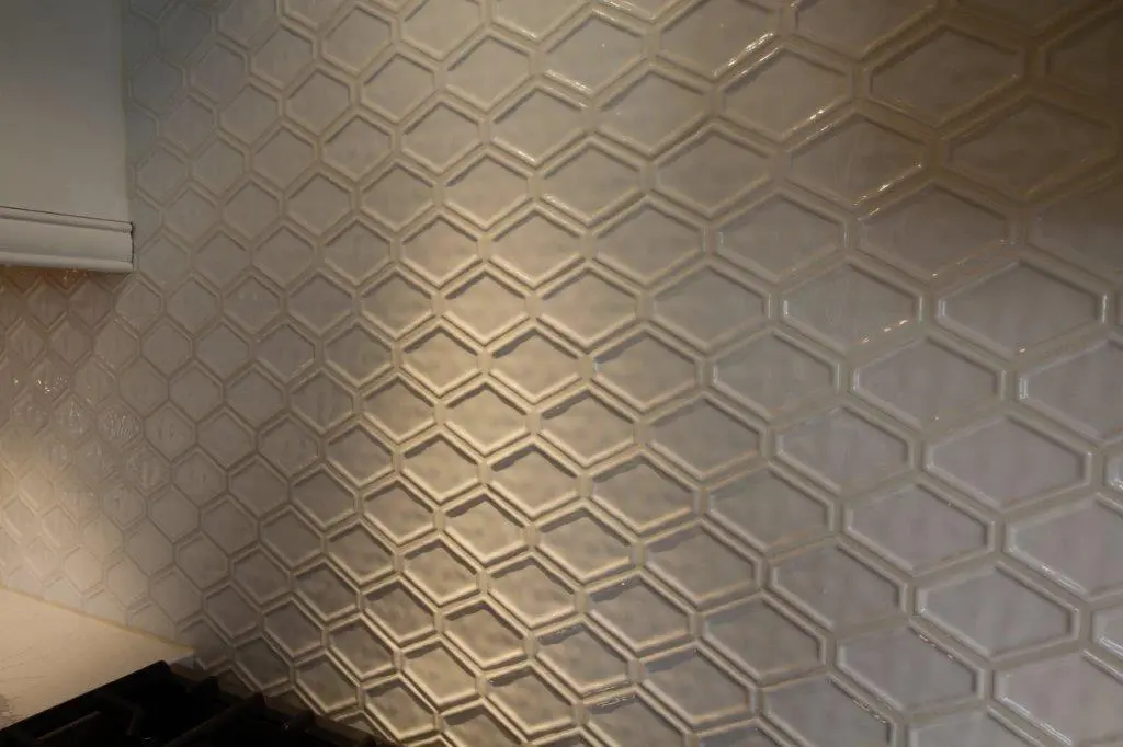 Close-up view of a kitchen backsplash with glossy, hexagonal and elongated hexagon white tiles arranged in a honeycomb pattern above a black stovetop and light-colored countertop.