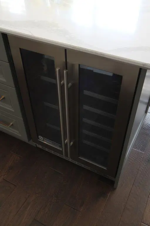 Under-counter dual glass-door beverage cooler with stainless steel handles, set in a dark wood floor kitchen with light-colored cabinetry and marble countertop.