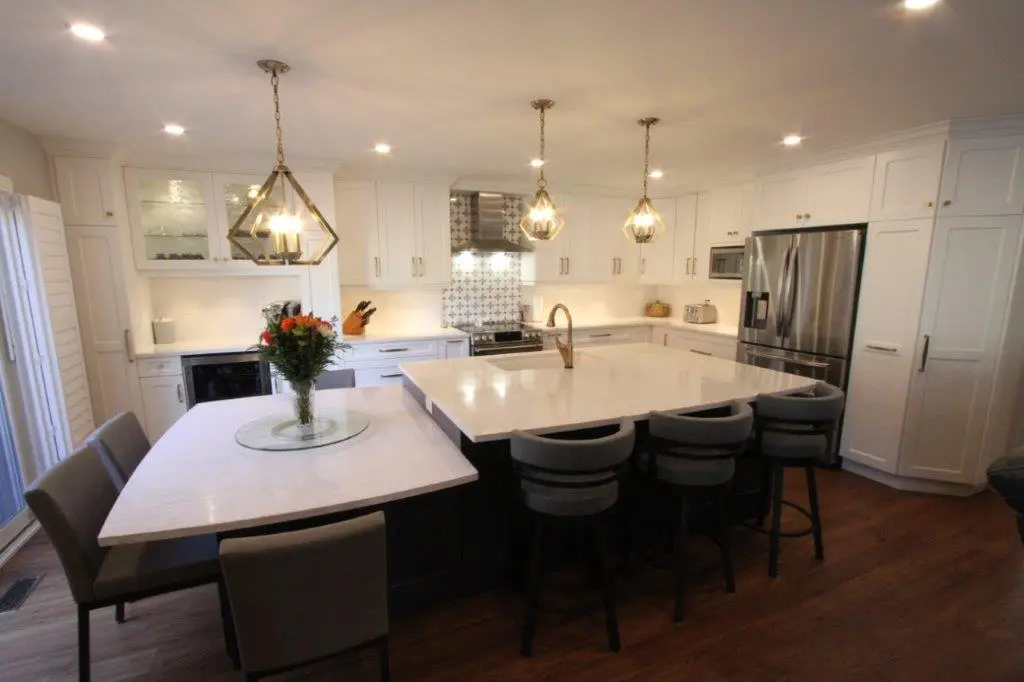 Modern kitchen with white cabinetry, a large island with four gray bar stools, a smaller attached table with four chairs, pendant lights above, stainless steel refrigerator, and a vase of flowers on the table.