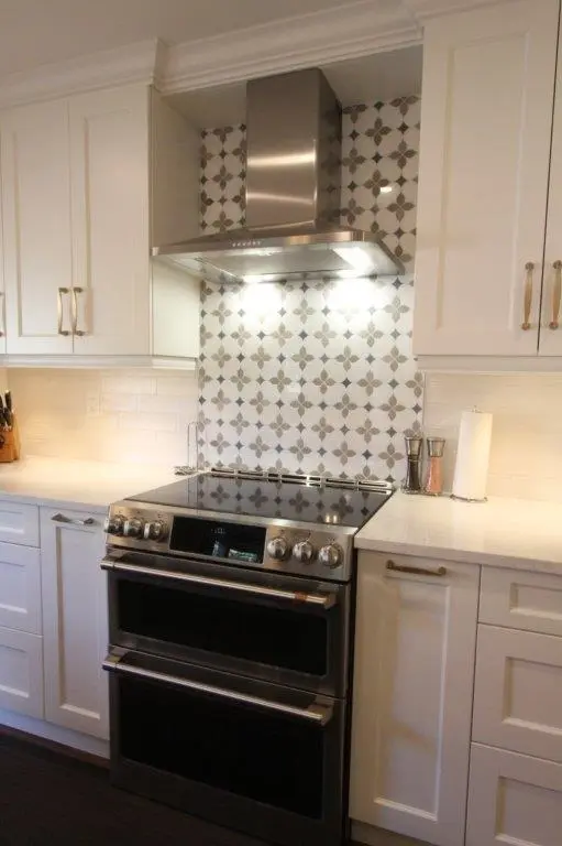 Modern kitchen with a stainless steel double oven and stovetop beneath a stainless steel range hood with patterned tile backsplash, surrounded by white cabinetry and light-colored countertops with pepper grinders and a paper towel roll on the right.