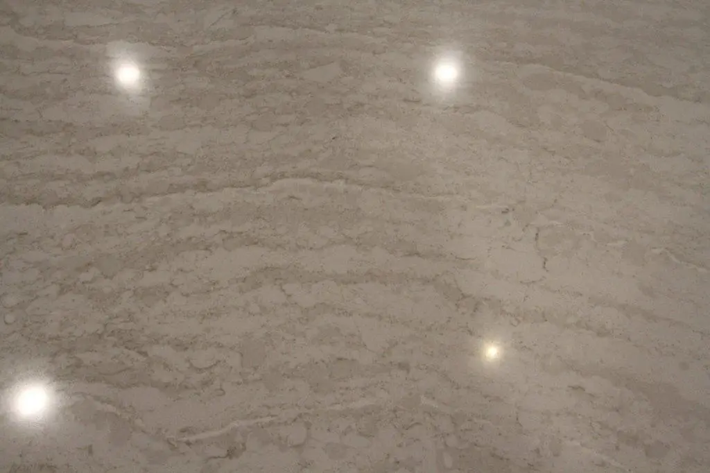 Reflective beige quartz surface with subtle natural veining and light spots reflecting overhead lights