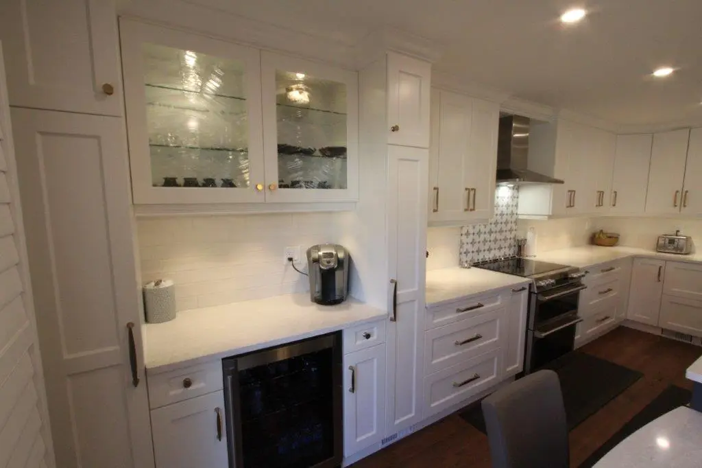 Modern kitchen with white cabinets, stainless steel appliances including a stove and wine cooler, white countertops, a coffee maker, and wood flooring under recessed ceiling lights.