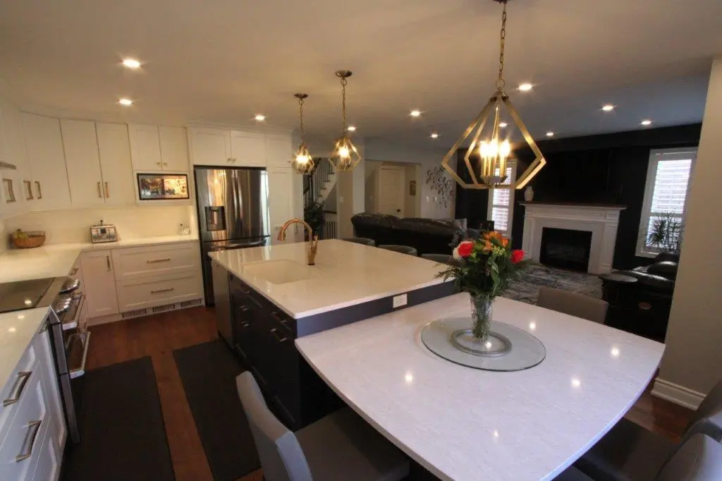 Modern open kitchen with white upper cabinets, dark lower island with white countertop, stainless steel refrigerator, wooden floor, and a dining table with a glass tray and a vase of colorful flowers in the foreground, leading to a living room with a fireplace, leather sofa, and dark wall accents.