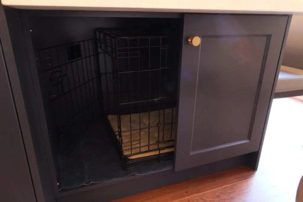 Black wire dog crate with a tan pad inside, placed within a dark cabinet with one door partially open, on a wooden floor.