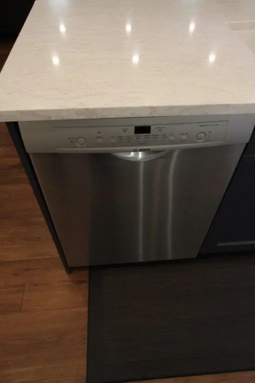 Stainless steel Bosch dishwasher installed under a light-colored quartz countertop in a kitchen with wooden flooring and a dark floor mat in front of it.