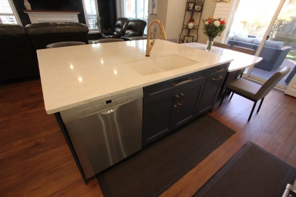 Modern kitchen island with a white quartz countertop, integrated sink with a gold faucet, dark cabinetry, and a stainless steel dishwasher, adjacent to a small dining table with chairs, set on wooden flooring with a view of a living area and glass doors leading outside.