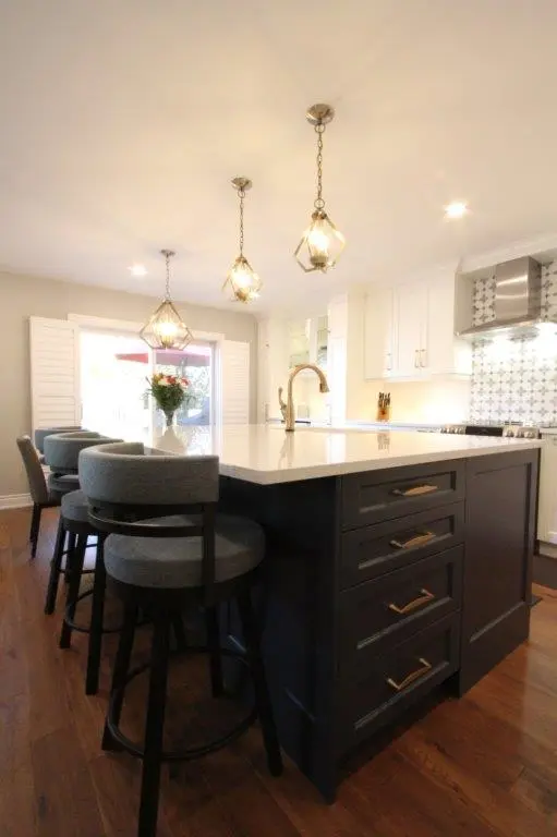 Modern kitchen with a large dark island featuring white countertop, four gray cushioned bar stools, three pendant lights, white cabinets, stainless steel range hood, and patterned backsplash tiles.