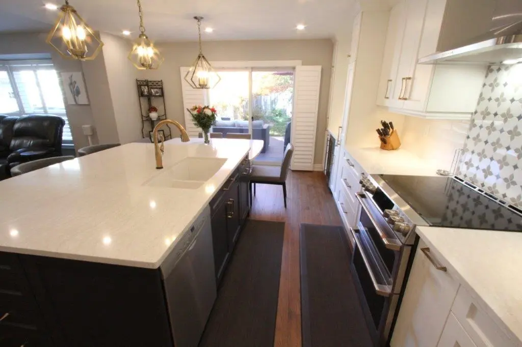 Modern kitchen with large white quartz island featuring a built-in sink and bronze faucet, three pendant lights overhead, dark lower cabinets, white upper cabinets, stainless steel appliances, wooden floor, and a sliding glass door leading to an outdoor patio.