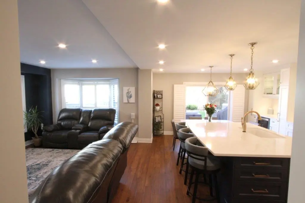 Open-concept living room and kitchen area with dark leather sofas, hardwood flooring, a large kitchen island with bar stools, pendant lights, a vase of flowers on the island, and large windows allowing natural light in.