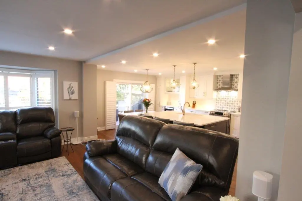 Open living room and kitchen with black leather sofas, a patterned area rug, wooden floors, and a kitchen island with pendant lights and a vase of flowers.