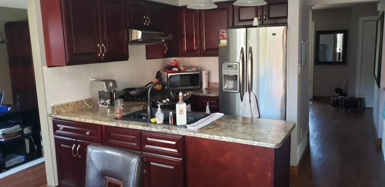 A before renovation picture of a Kitchen with dark wood cabinets and a granite countertop island featuring a black faucet, soap dispenser, and bottles, a stainless steel refrigerator, microwave, stove with a pan, and an espresso machine, with a hallway visible to the right.