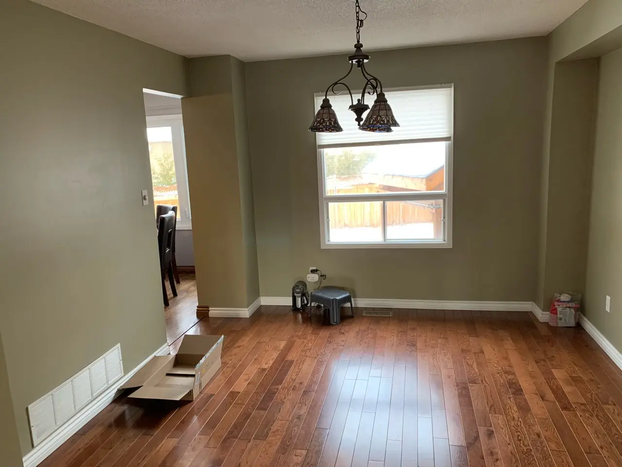 A before renovation picture of an Empty room with brown wooden floor and olive green walls, featuring a window with a white blind, a hanging chandelier light fixture, an open cardboard box on the floor near a vent, a small gray stool, and some items in the corner on the floor near the wall outlet.