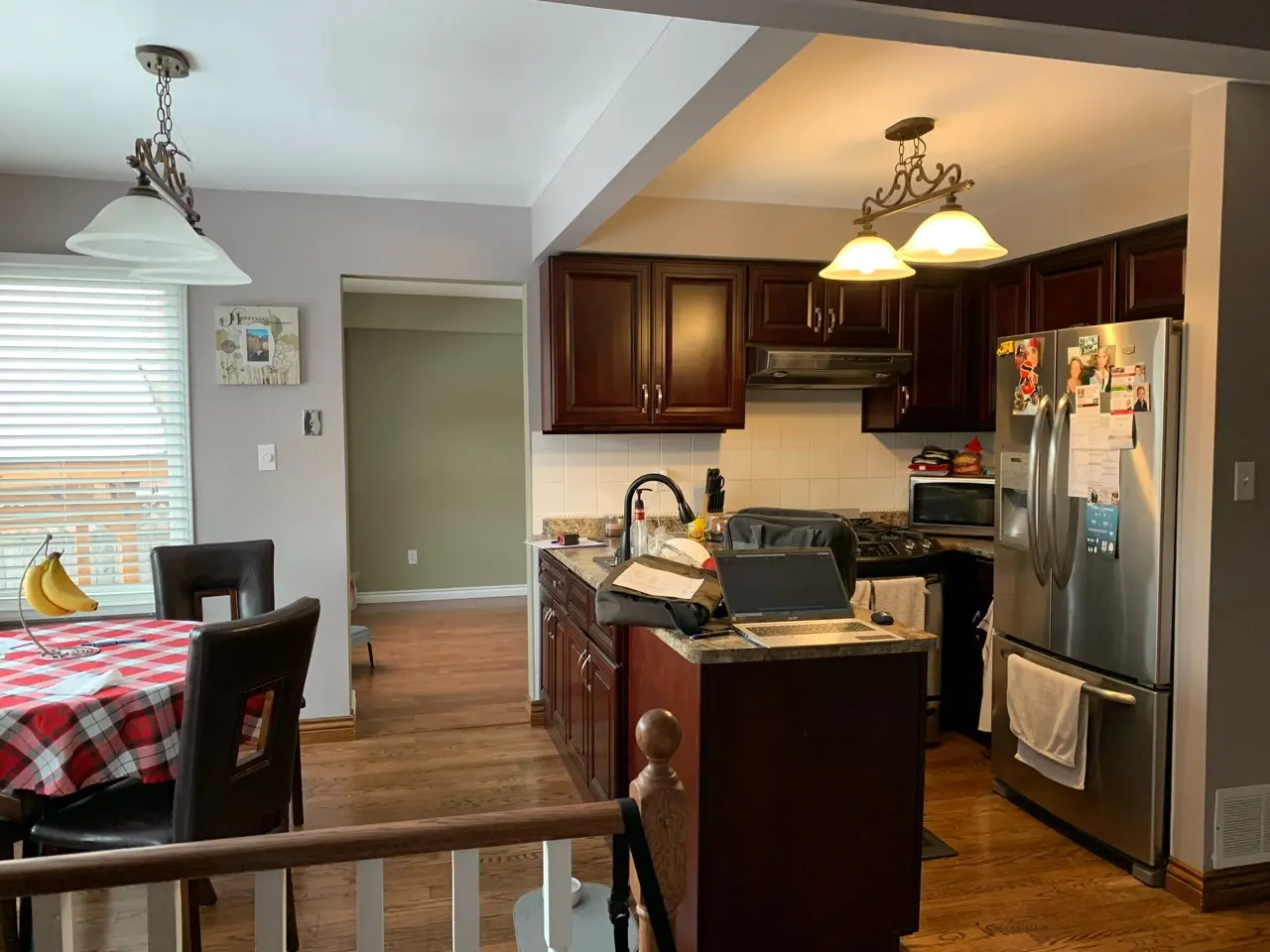 A before renovation View of a kitchen and dining area featuring dark wood cabinets, stainless steel refrigerator with magnets and notes, a kitchen island with laptop and bags, a dining table with a red and white checkered tablecloth and hanging banana holder, hardwood floors, and pendant lighting above both spaces.