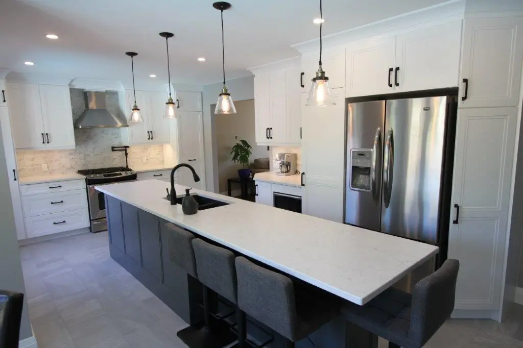 Modern kitchen with a long white quartz island featuring a black sink and faucet, four gray cushioned bar stools, white upper cabinets, stainless steel refrigerator and oven, and three pendant lights hanging above the island.