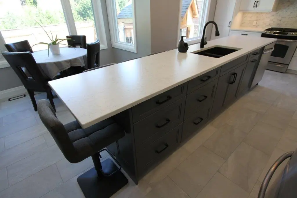 Modern kitchen island with white countertop, dark gray cabinetry with black handles, a black sink and faucet, and a gray upholstered bar stool, next to a dining area with a round table covered by a white tablecloth and black chairs, with large windows in the background.