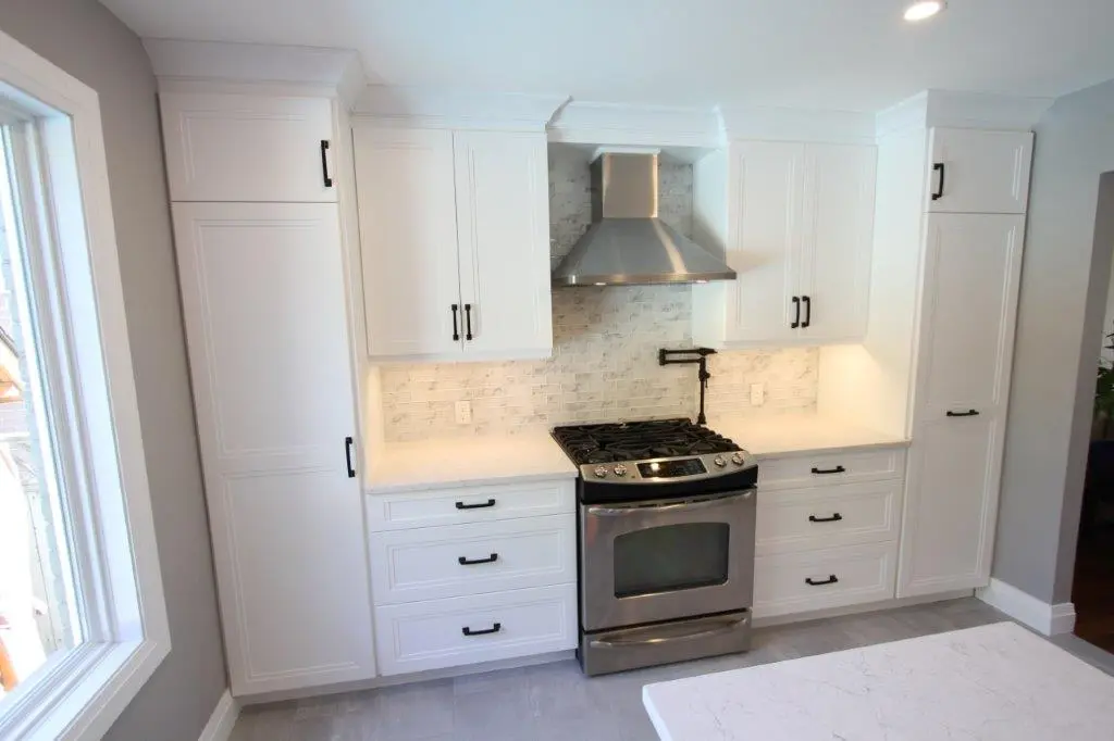 Modern kitchen with white cabinets, a stainless steel gas stove and range hood, white quartz countertops, a gray brick backsplash, and a large window on the left side.