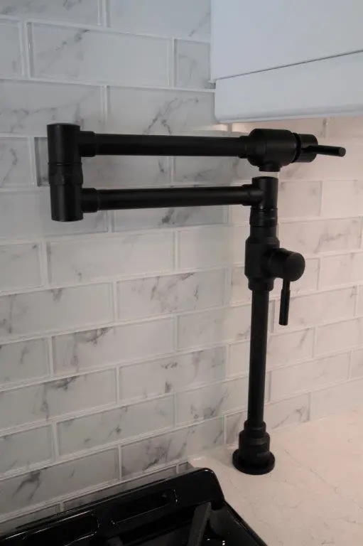 Matte black pot filler faucet with dual-arm joints mounted on a white marble-patterned subway tile backsplash above a countertop and stove.