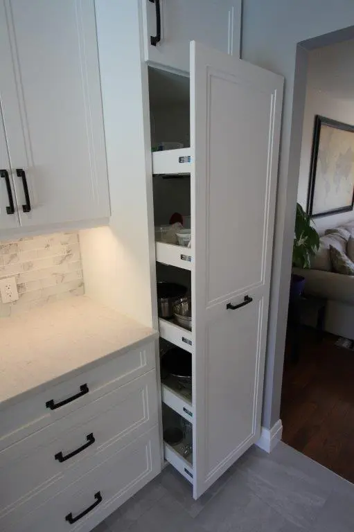 Tall white kitchen cabinet with a partially open door revealing pull-out shelves holding pots, pans, and containers, next to a countertop with white drawers and black handles, with a view into a living area in the background.